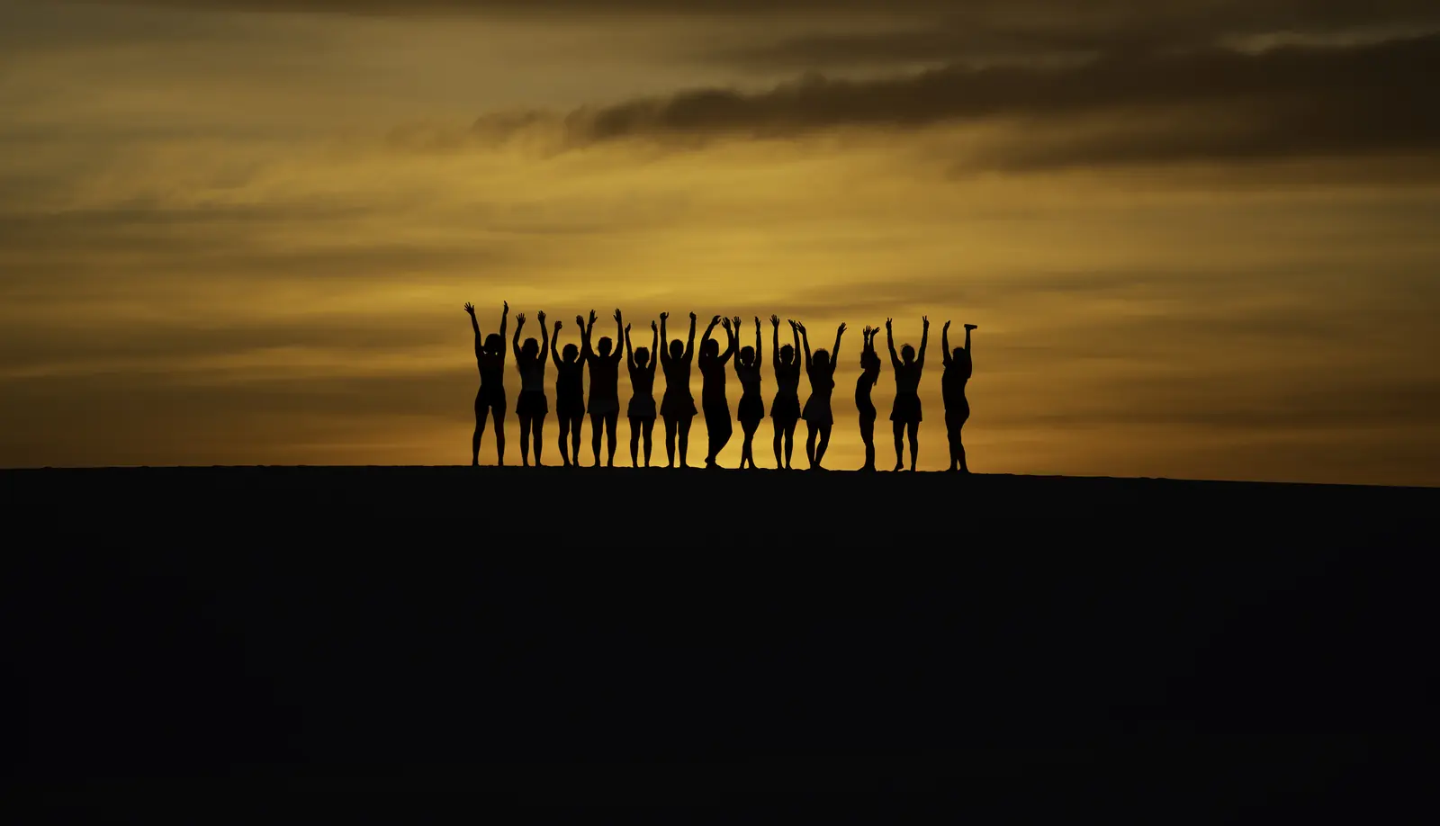 Grupo de braços erguidos no topo de uma duna ao pôr do sol, na travessia dos Lençóis Maranhenses