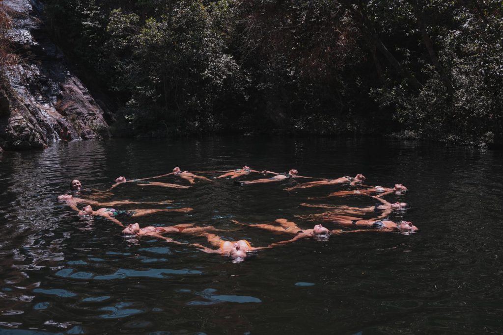 Mulheres boiando de mãos dadas em roda no poço de uma cachoeira cercada de mata